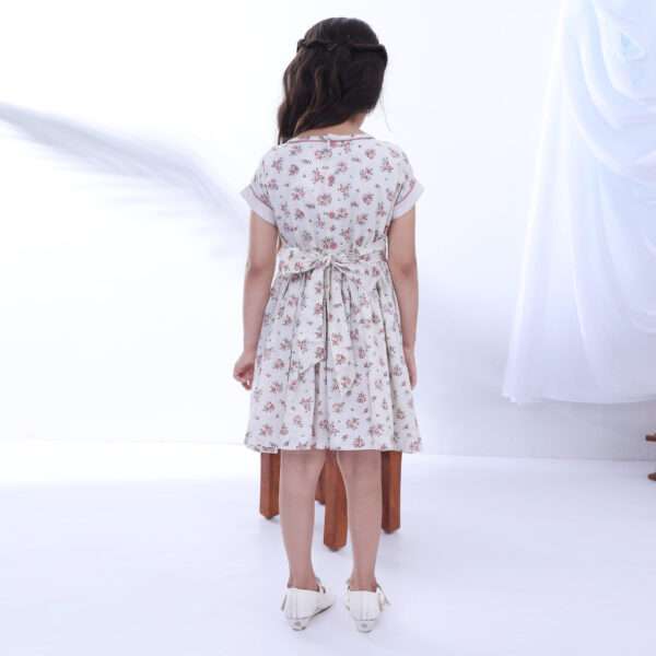Rear image of a girl wearing floral printed ivory cotton dress with back tie-up sash and contrast cord piping details on the collar and sleeves