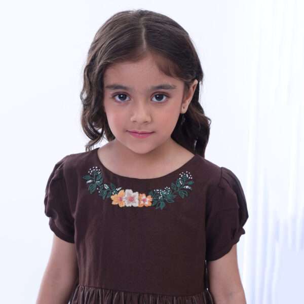 Girl looks at camera with a half smile, she is dressed in a brown dress with gathered sleeves and floral embroidered neckline