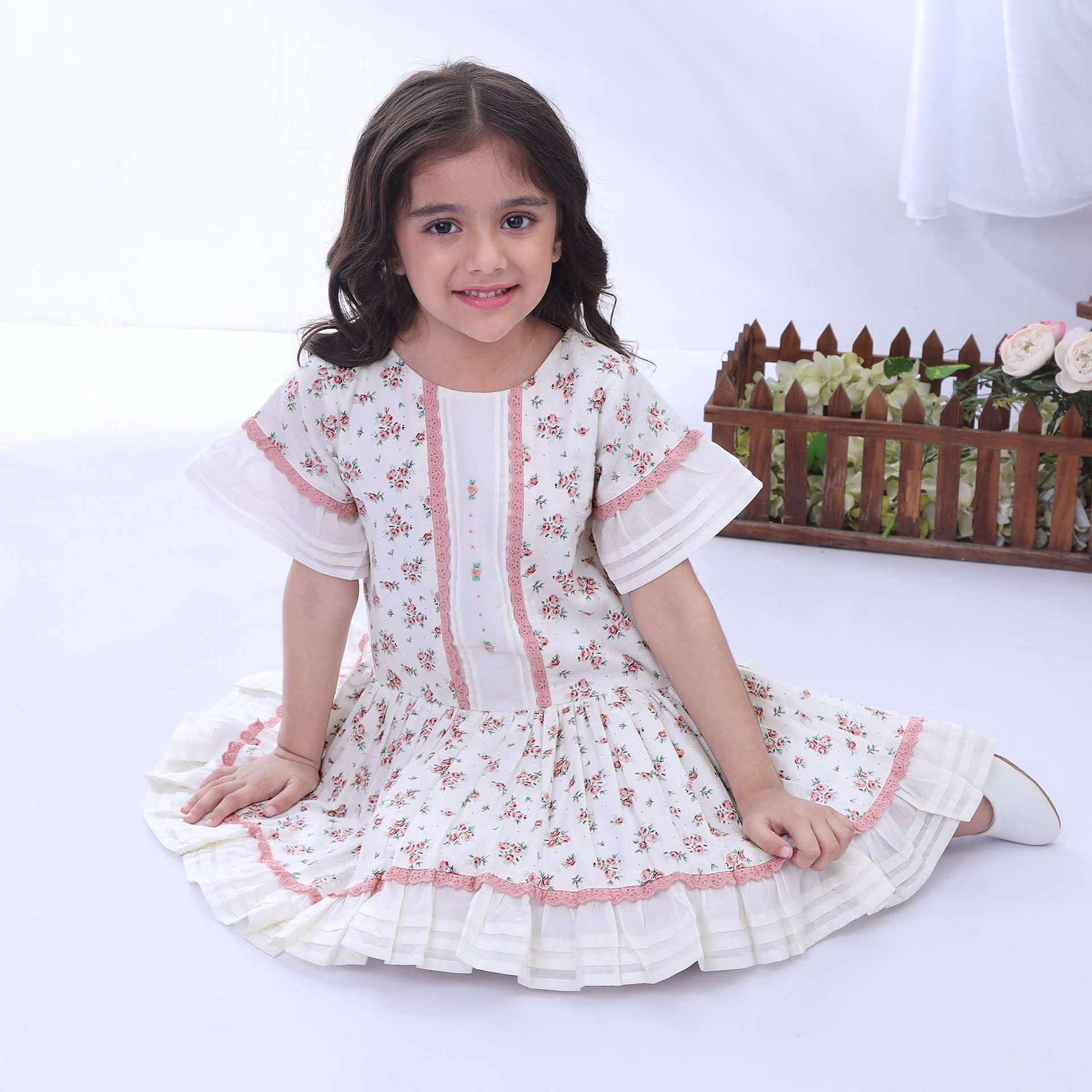 A seated girl in ivory floral printed dress with hand-embroidery, dropwaist pattern with pleated hem and sleeve ruffles