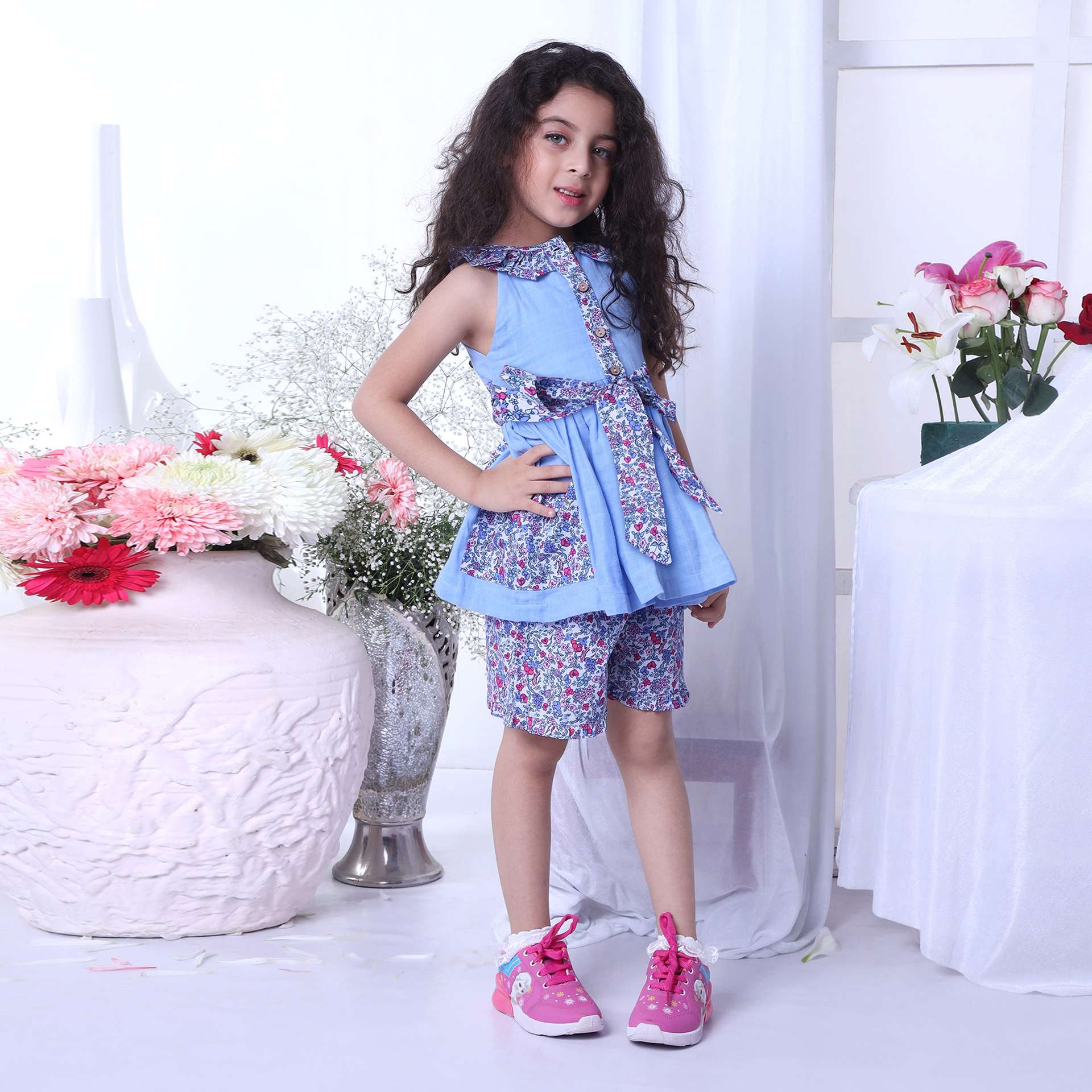 A young girl wearing blue gauze blouse with cutaway shoulders with floral trims, tie-up belt and matched floral shorts