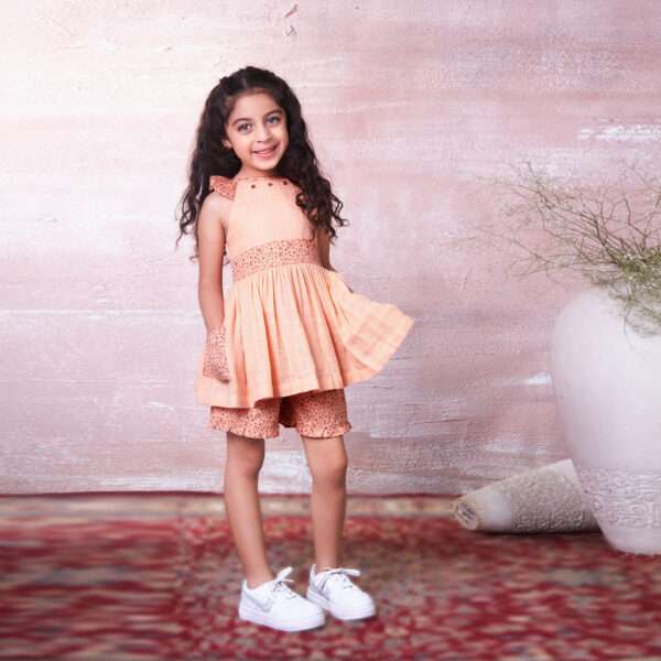 A little girl in peach gauze tunic with ruffles, hand embroidery and ties in a floral trim, with matching floral shorts