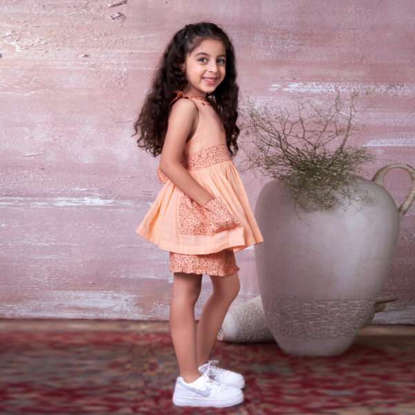 A little girl in peach gauze tunic with ruffles, hand embroidery and ties in a floral trim, with matching floral shorts