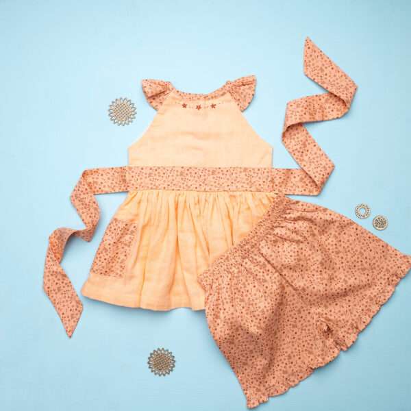 Flatlay image of peach gauze tunic with ruffles, hand embroidery and ties in a floral trim, with matching floral shorts