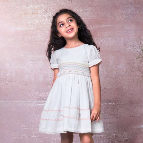 A little girl wearing a delicately hand-smocked cotton dress with ric-rac trims and pastel floral embroidery