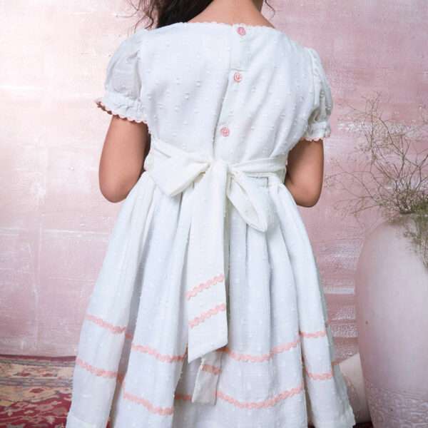 Rear view of a little girl wearing a delicately hand-smocked cotton dress with ric-rac trims and floral embroidery