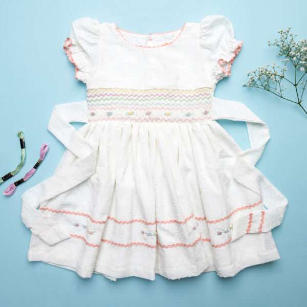 Flatlay image of delicately hand-smocked cotton dress with ric-rac trims and pastel floral embroidery