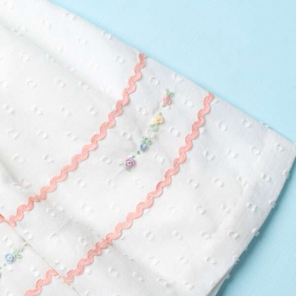 Close-up image of a flatlay of the hem of a hand-smocked cotton dress with ric-rac trims and pastel floral embroidery