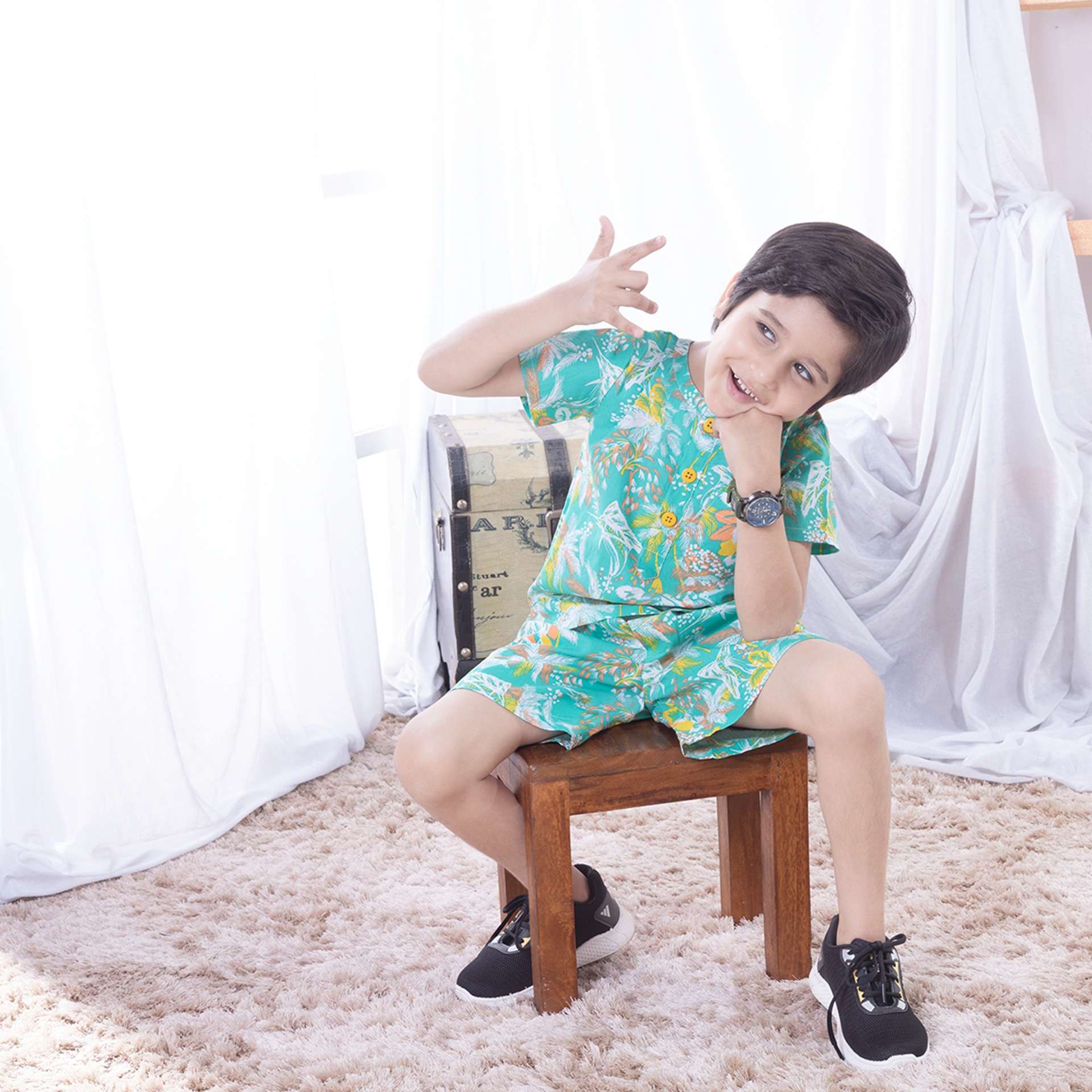 A little boy seated on a stool posing in green tropical palm printed cotton coordinated shirt and shorts set