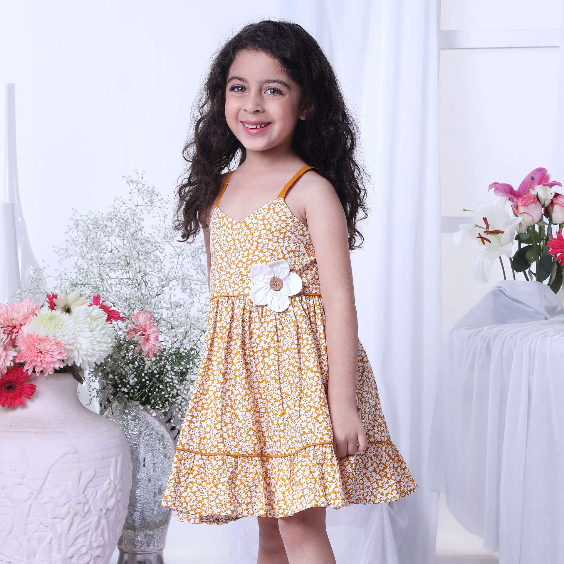 A little girl wearing strappy shoulder dress with floral print in mustard and big flower appliqued on the waist
