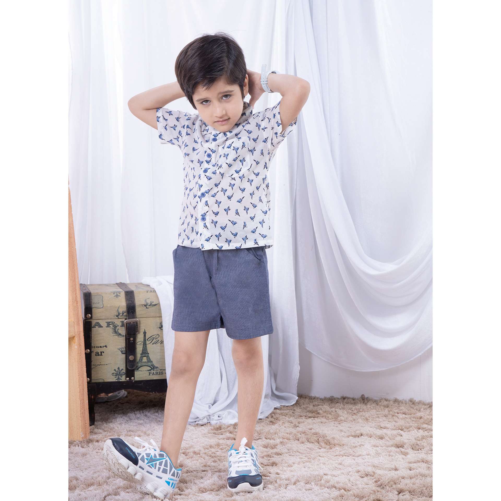 A little boy in white bird print shirt with buttons down the front paired with grey shorts