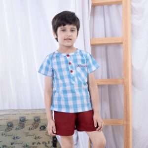 Small boy standing dressed in a blue plaid shirt with embroidered pocket and deep red shorts