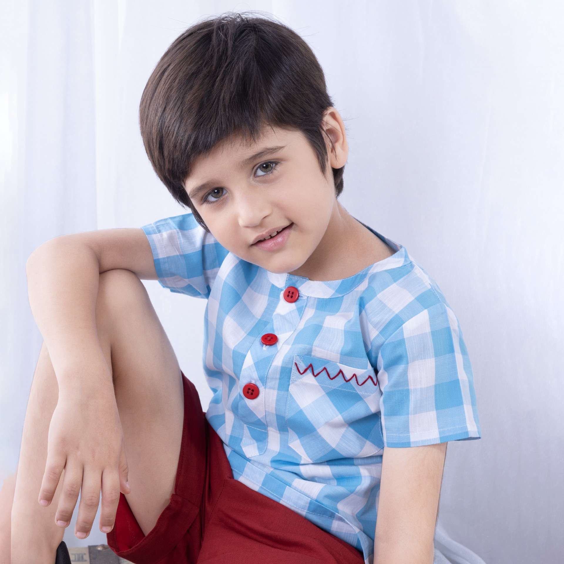 A little boy in light blue checked shirt with red hand embroidered pocket and red buttons paired with matching shorts