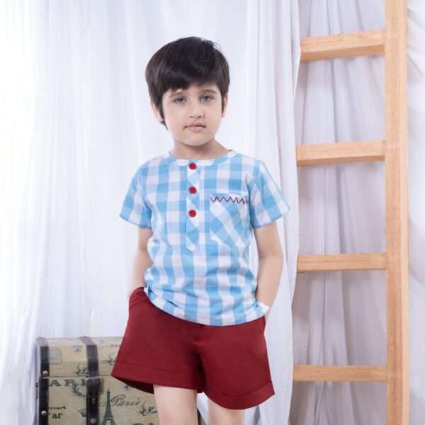 Boy stands in a casual stance wearing a blue big check collarless shirt and bright red shorts with side pockets