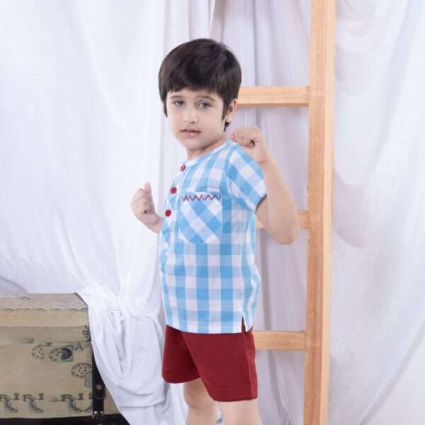 A little boy in light blue checked shirt with red hand embroidered pocket and red buttons paired with matching shorts