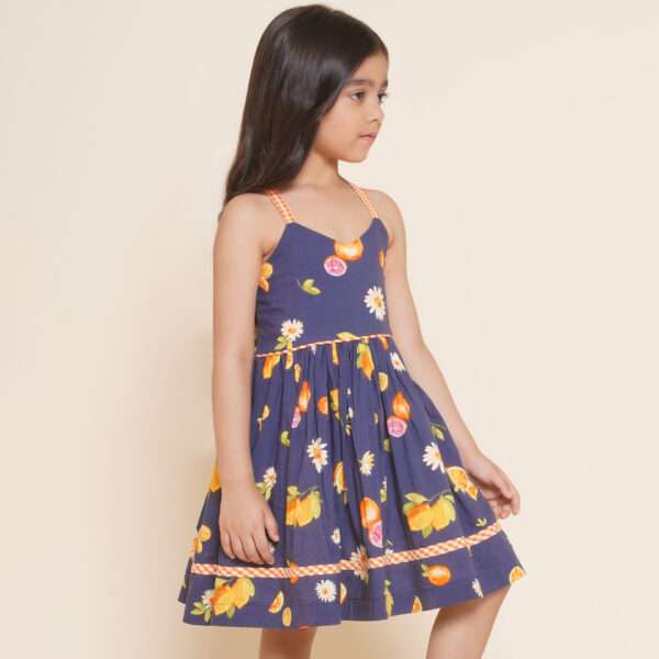 A little girl wearing strappy vibrant fruit printed dress with orange gingham trims, shirred back and tie-up straps