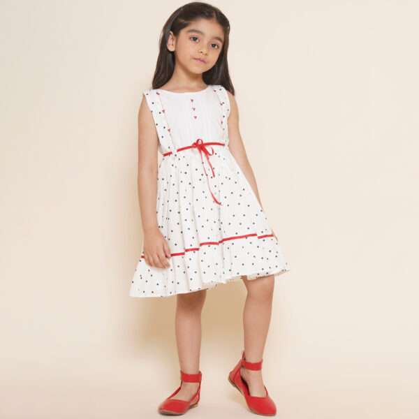 A little girl in sleeveless polka dot ivory cotton dress with red satin belt and hand embroidered bow embellishments
