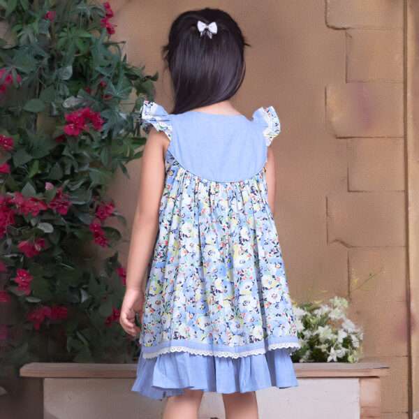 Rear image of a girl in blue chambray floral printed cotton dress with flutter sleeves, eyelet lace trims and ruffles