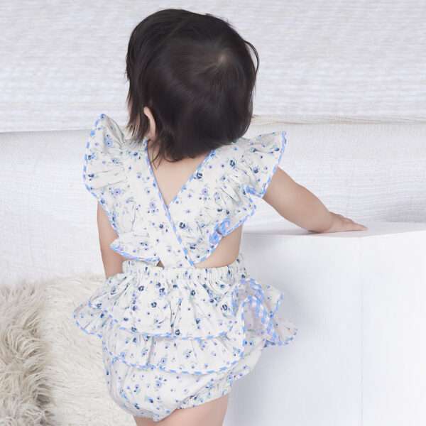 Rear image of baby girl in blue floral printed onesie with back crossover straps and ruffles on the bottom