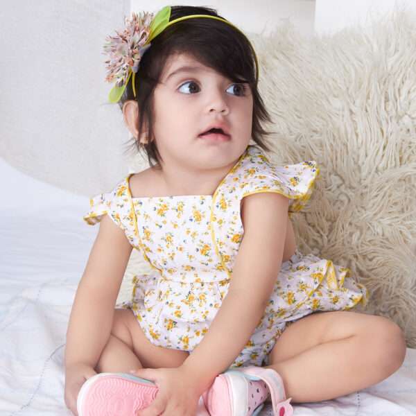 A baby girl in yellow floral printed onesie with back crossover straps and ruffles on the bottom with contrast trims