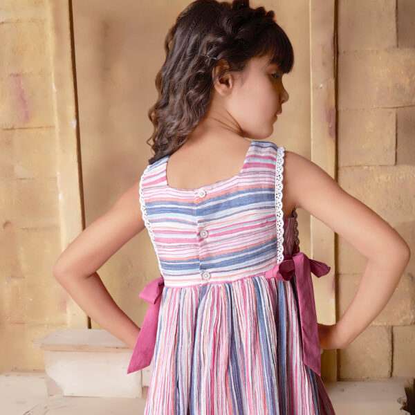 Rear side of a little girl in sleeveless multi-color stripe dress with side ties, lace trims and a decorative thread bow