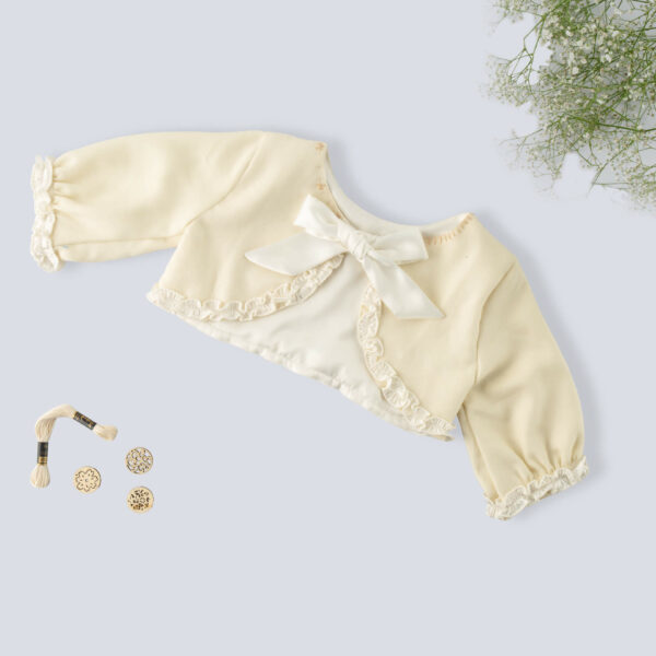 Flat lay view of an ivory bolero embroidered jacket for girls featuring a tie-up sash bow with props