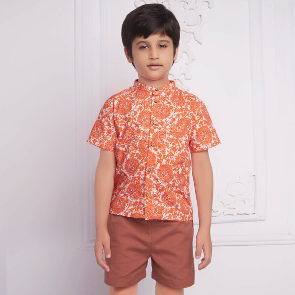 Boy wears an orange floral printed mandarin collared shirt matched with rust shorts, both with pockets.