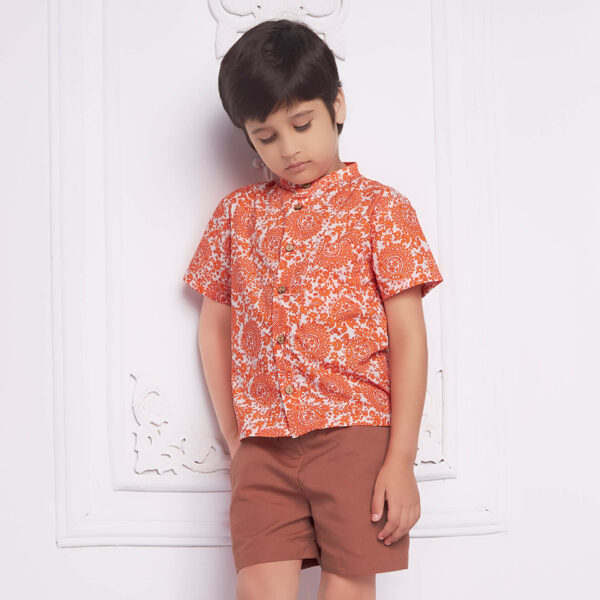 Boy leans against a wall wearing an orange indian print shirt and rust tone shorts