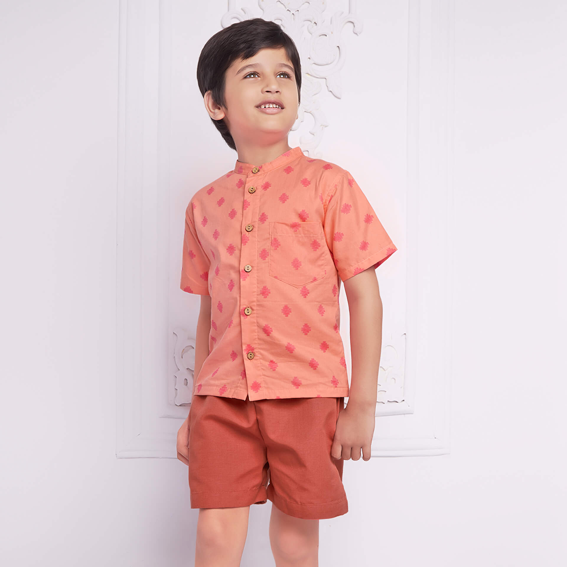 Boy wears peach printed shirt matched with rust shorts, both with pockets.