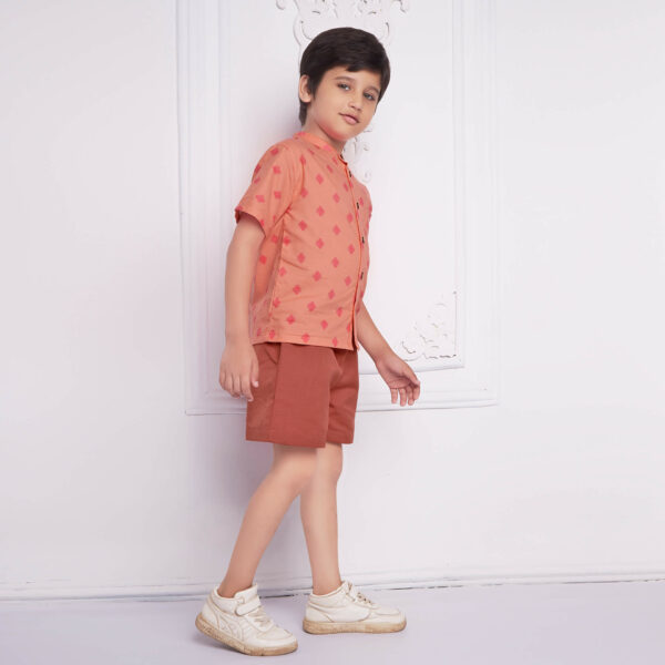 A boy strikes a walking pose, in a peach printed mandarin collared shirt and rust shorts.