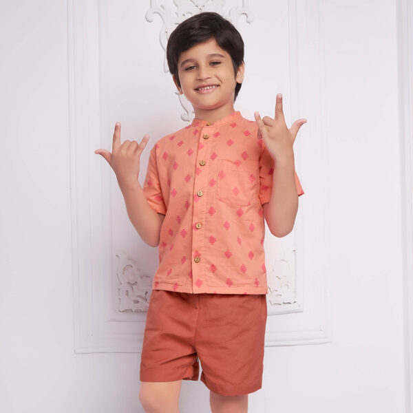 A boy strikes a pose, in a peach printed mandarin collared shirt and rust shorts.