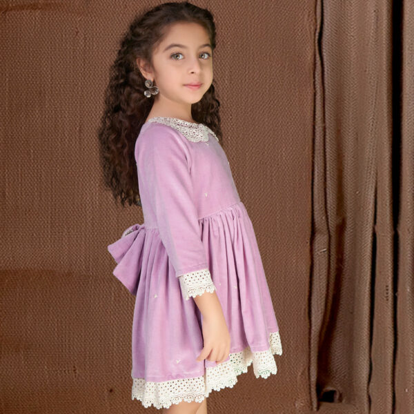 Side pose of a girl wearing an embroidered lavender velvet dress with an attached embellished bow at the waist with lace trims