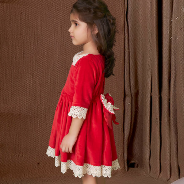 From the side, a girl showcases red velvet dress adorned with ivory lace and a large bow at the back