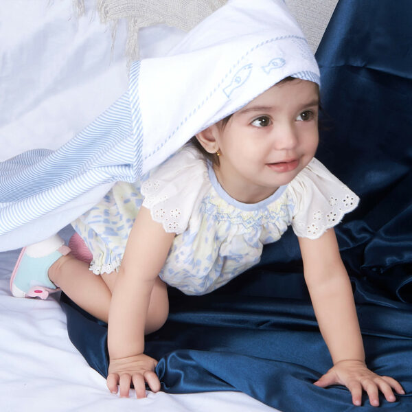 A baby girl crawls in a watercolor smocked onesie adorned with lace trims, wearing a "Something Fishy" blanket as a cap towel on her head