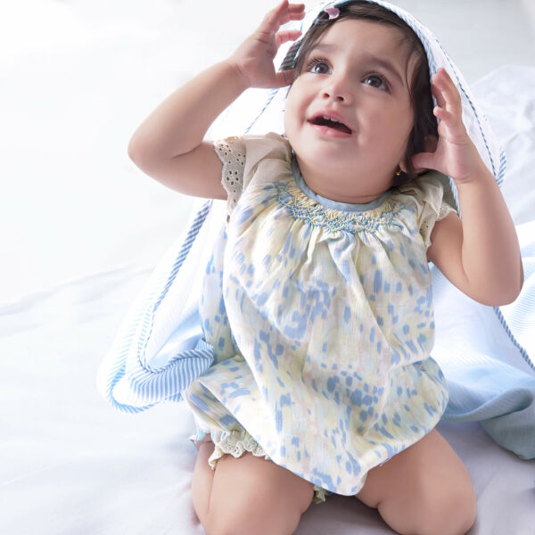 A baby girl in a watercolor smocked onesie adorned with lace trims, wearing a "Something Fishy" blanket as a cap towel on her head