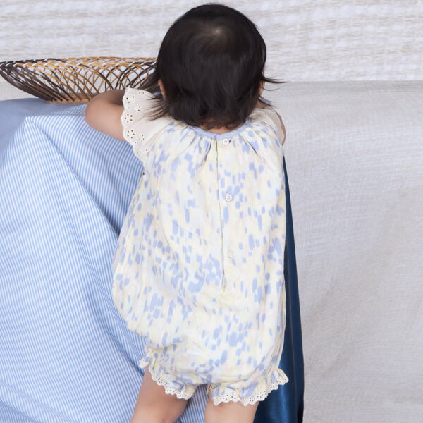 Back view of a baby girl in a watercolor smocked onesie adorned with lace trims, fastened by back buttons.