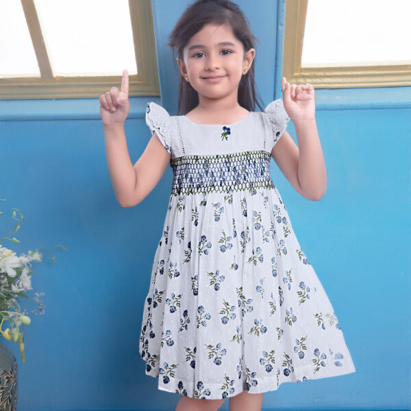 A girl poses with both hands up, wearing a white swiss dot floral printed smock dress