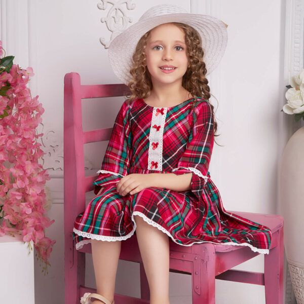 Seated with a hat atop her head, a little girl wears red plaid dress with white lace and red satin bows as trims