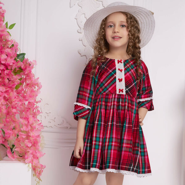 With a hat atop her head, a little girl wears red plaid dress with white lace and red satin bows as trims