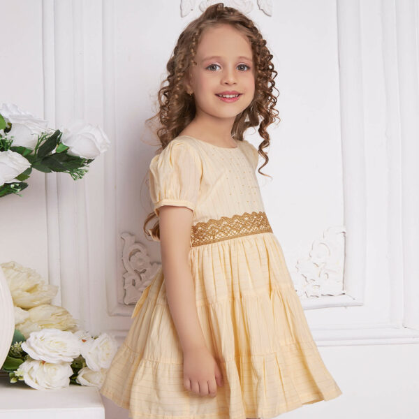 Posing from the side, a little one wears an ivory beads dress with gold metallic lace at the waist
