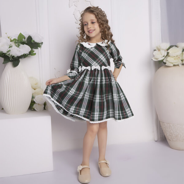Elegantly posing, a young girl showcases a green tartan collar embroidered dress with bows at the waist