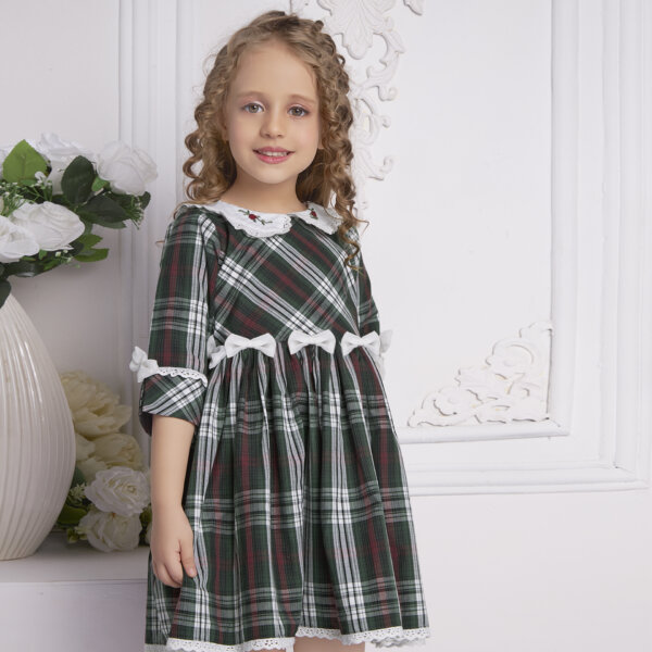 A young girl showcases a green tartan collar embroidered dress with bows at the waist in a slightly turned side view