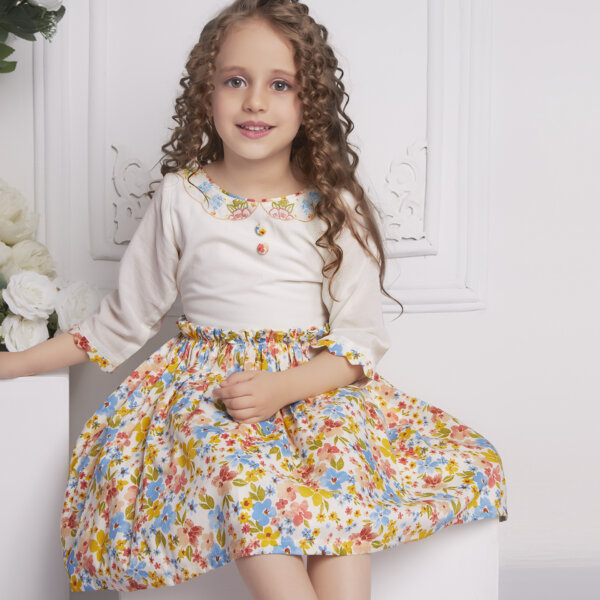 A little girl seated wearing ivory floral dress with delicate floral hand-embroidered neckline