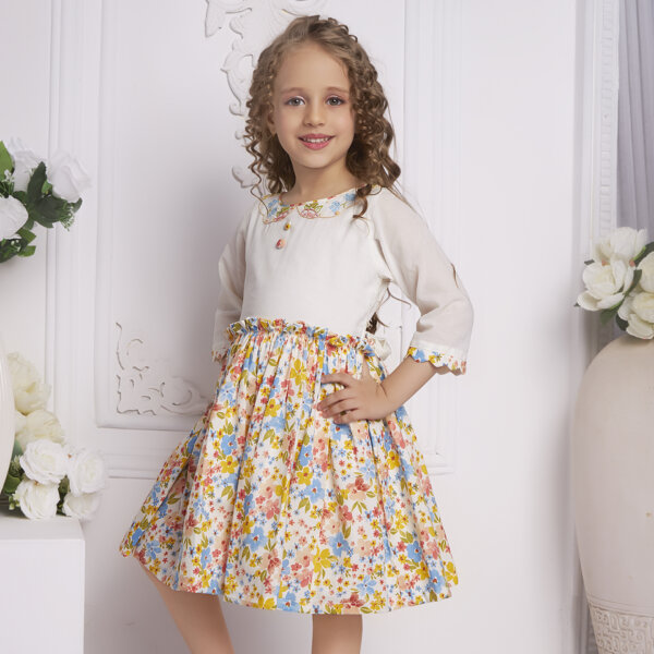 With hands on hips, a side pose of a little girl in an ivory floral embroidered dress with decorative cloth buttons