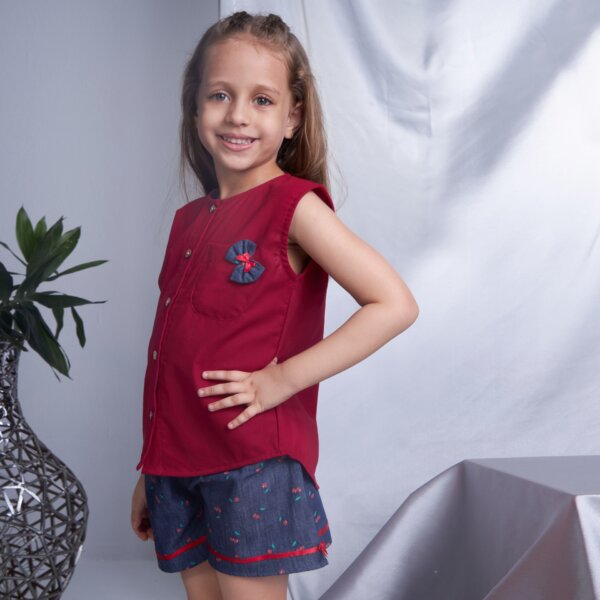 Side pose of a little girl wearing sleeveless red blouse and navy blue cherry printed shorts, featuring a bow detail on the pocket.