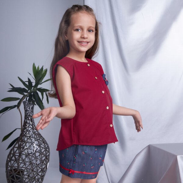 Side pose of a little girl wearing sleeveless red blouse and navy blue cherry printed shorts