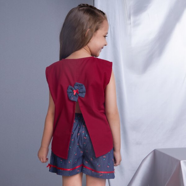 A girl is seen from the back, wearing a red sleeveless blouse with a slit at the back, paired with cherry printed navy shorts featuring a satin ribbon trim.