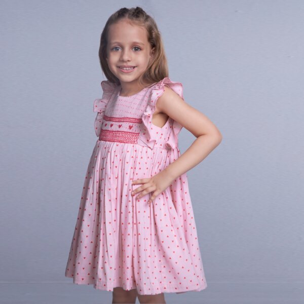 With hands on hips, a girl wears a pink cotton dress with heart embroidery and smock, posing with a smile