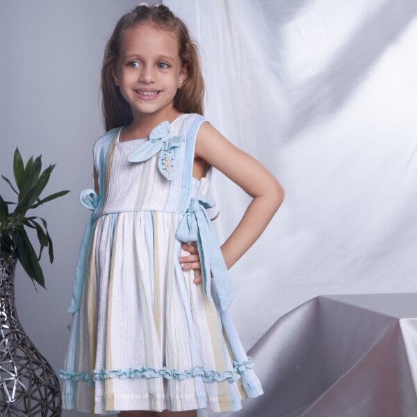 A little girl in sleeveless blue lurex embroidered dress with side ties, with her hand on her hip and a side view