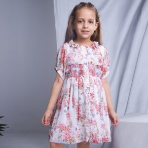 In an ivory floral smock dress, a child stands with hands straight, the collar adorned with ruffles