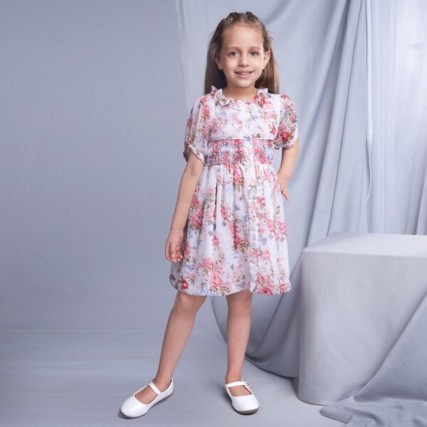 A little girl wears an ivory floral printed smock dress with decorative front fastened buttons, featuring ruffles on the collar